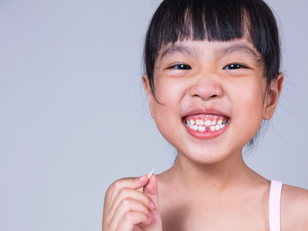 Young Asian girl with one missing tooth and holding the tooth that fell out.